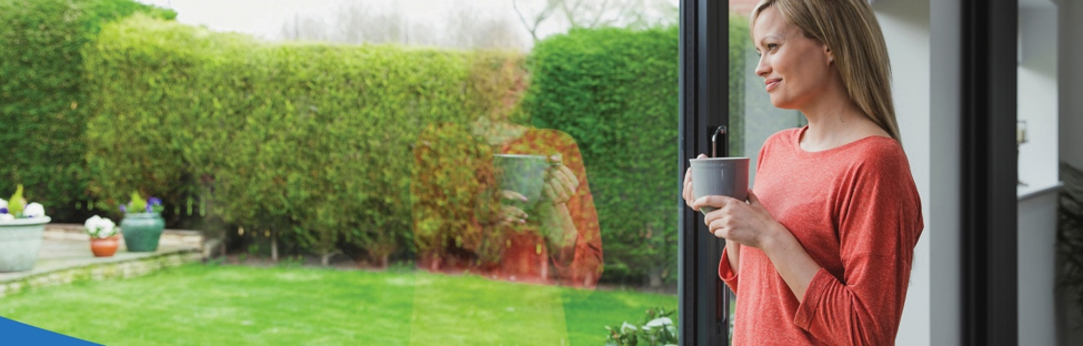 Blonde woman holding mug looking happily out her window at her green grass in the yard