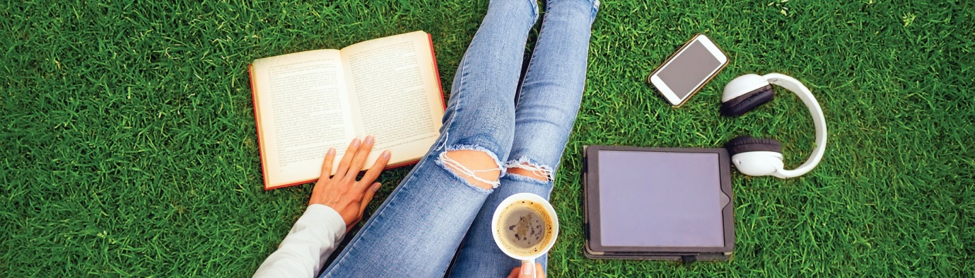 woman sitting on green grass with a book, mug, headphones, phone, and tablet